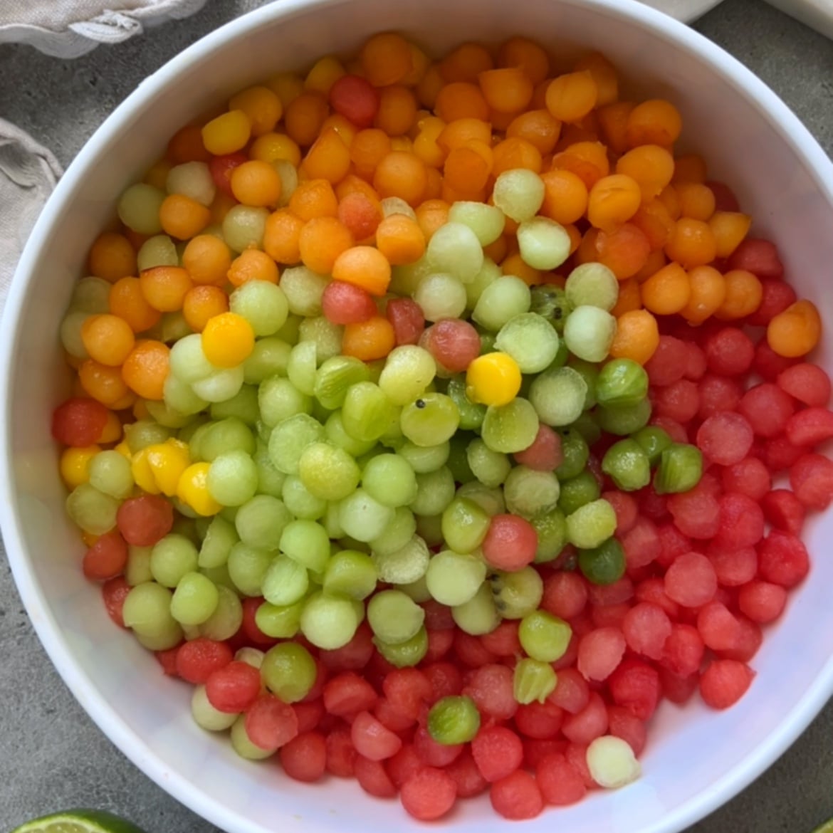 A white bowl filled with colorful melon balls, including apricot, green Galia melon, and red watermelon, arranged in sections.