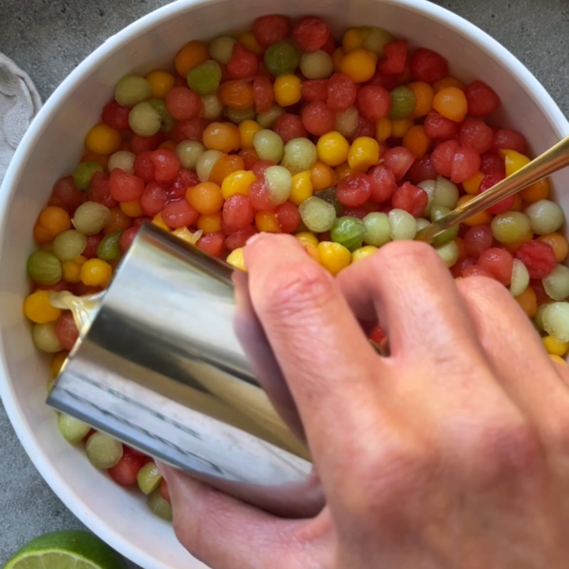 A hand pours lime dressing over a bowl of colorful melon balls.