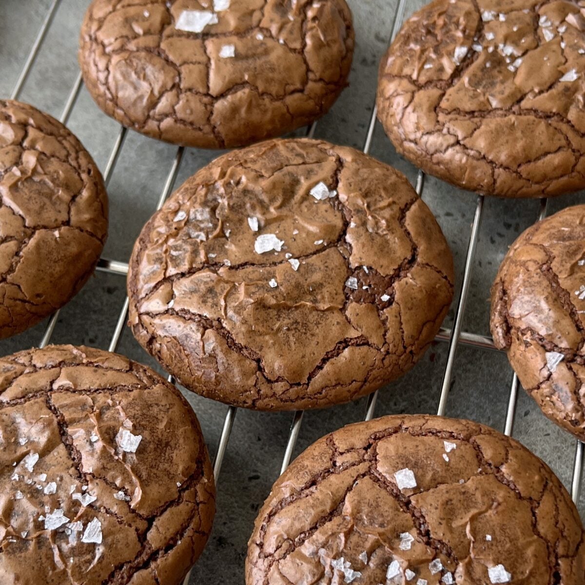 Several brownie cookies with cracked surfaces and flaky sea salt on top are cooling on a metal rack.