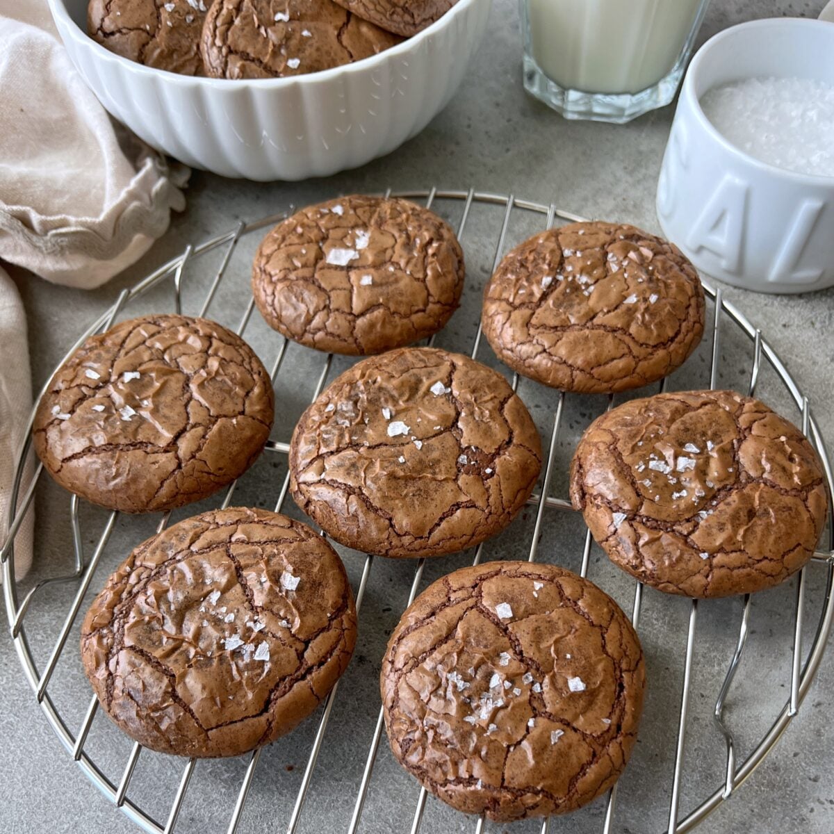 Seven rich brownie cookies sprinkled with flaky sea salt are cooling on a wire rack, with a bowl, a cup of milk, and a small dish of salt in the background.