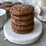 A stack of five brownie cookies sprinkled with sea salt sits on a round marble board. More cookies and a glass of milk are in the background.