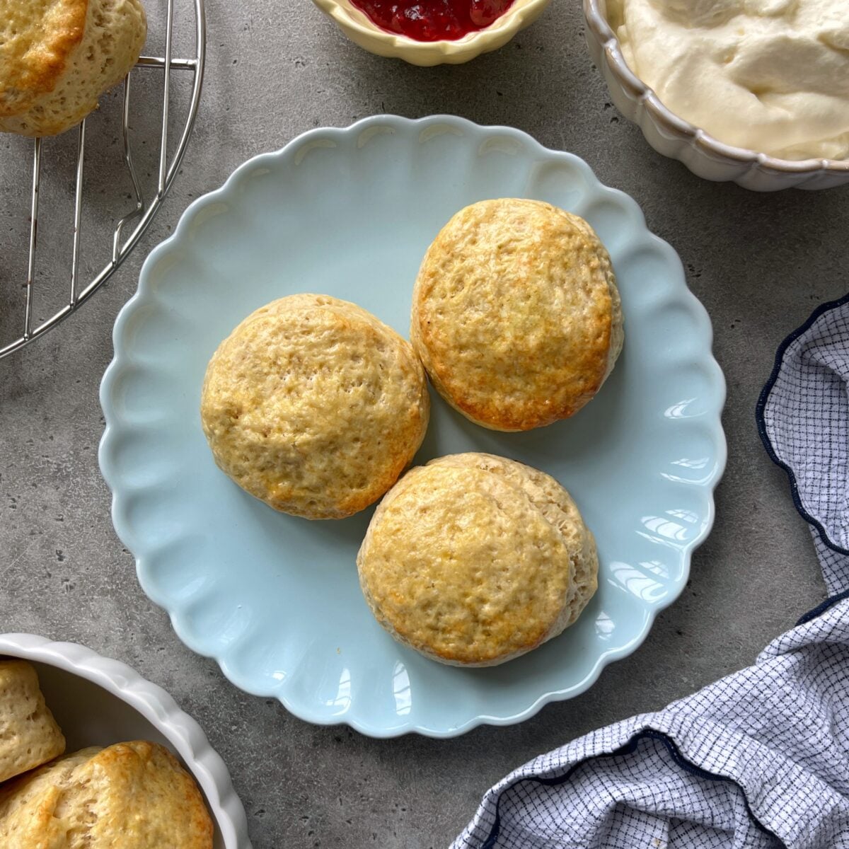 Three plain scones are arranged on a light blue scalloped plate, with cream, jam, and a cooling rack nearby.
