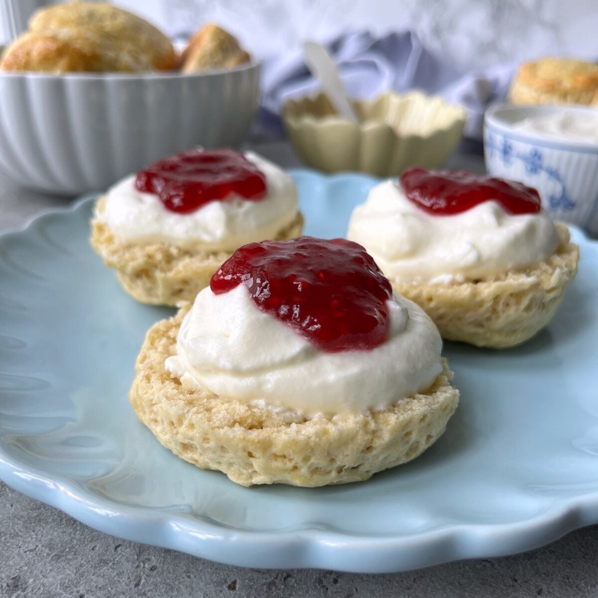 Three scones topped with whipped cream and red jam sit on a light blue plate.