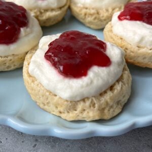 Round scones topped with a layer of whipped cream and a dollop of red jam are arranged on a light blue plate.
