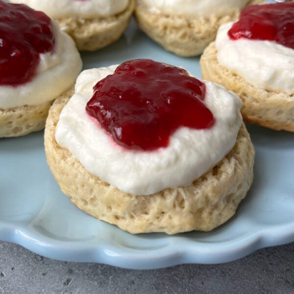 Round scones topped with a layer of whipped cream and a dollop of red jam are arranged on a light blue plate.