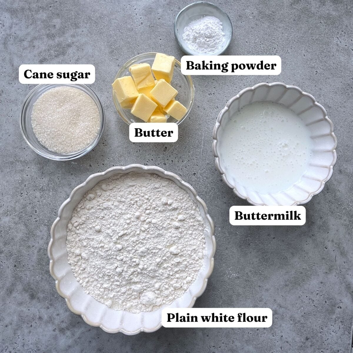 Five bowls containing cane sugar, butter, baking powder, buttermilk, and plain white flour are arranged on a gray surface. Each ingredient is clearly labeled.