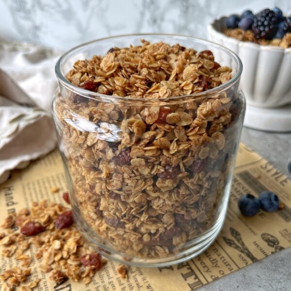 A glass jar filled with cinnamon granola sits on a piece of newspaper, with some granola spilled beside it. A bowl of yogurt, granola, and berries is in the background.
