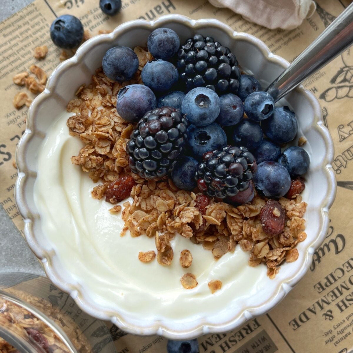 A bowl of yogurt topped with cinnamon granola, blueberries, and blackberries, with a spoon inside, sits on a table scattered with berries and a newspaper-style placemat.