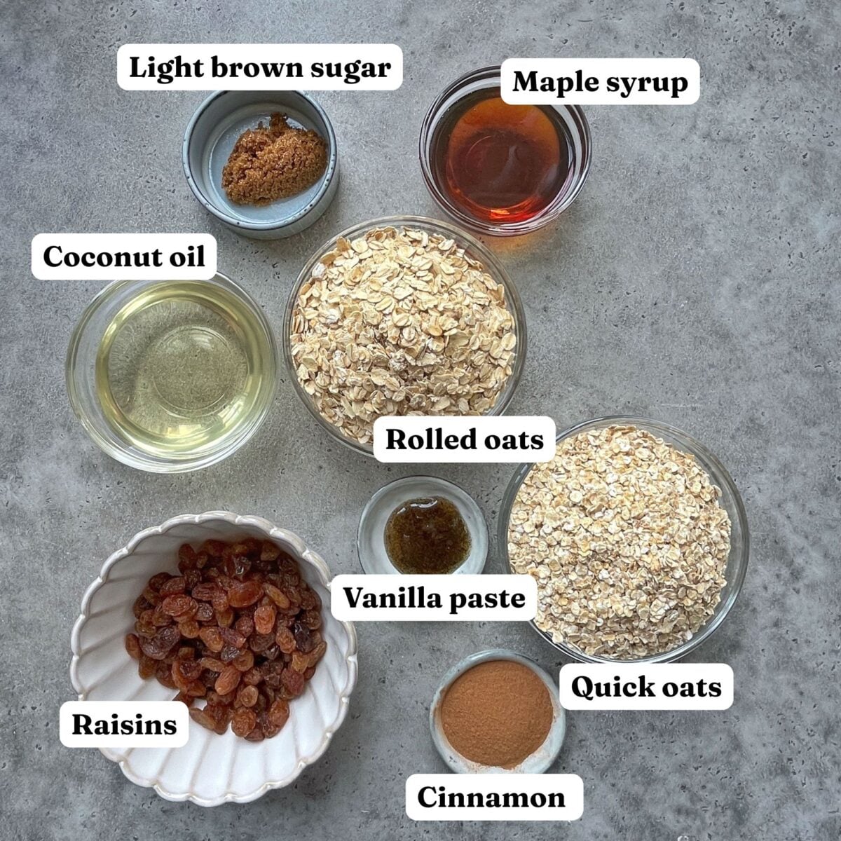 Assorted ingredients for Cinnamon Granola in bowls on a gray surface, including light brown sugar, maple syrup, coconut oil, rolled oats, quick oats, vanilla paste, raisins, and cinnamon. Labels identify each item.