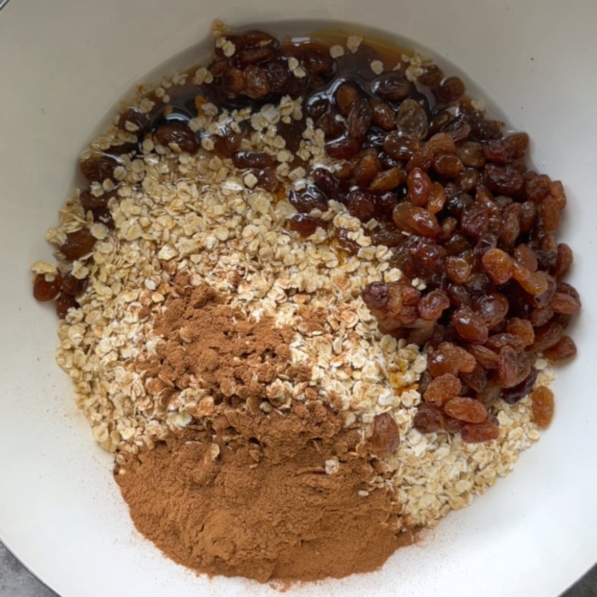 A mixing bowl containing oats, raisins, ground cinnamon, maple syrup, and cinnamon, arranged in separate sections before being mixed together.