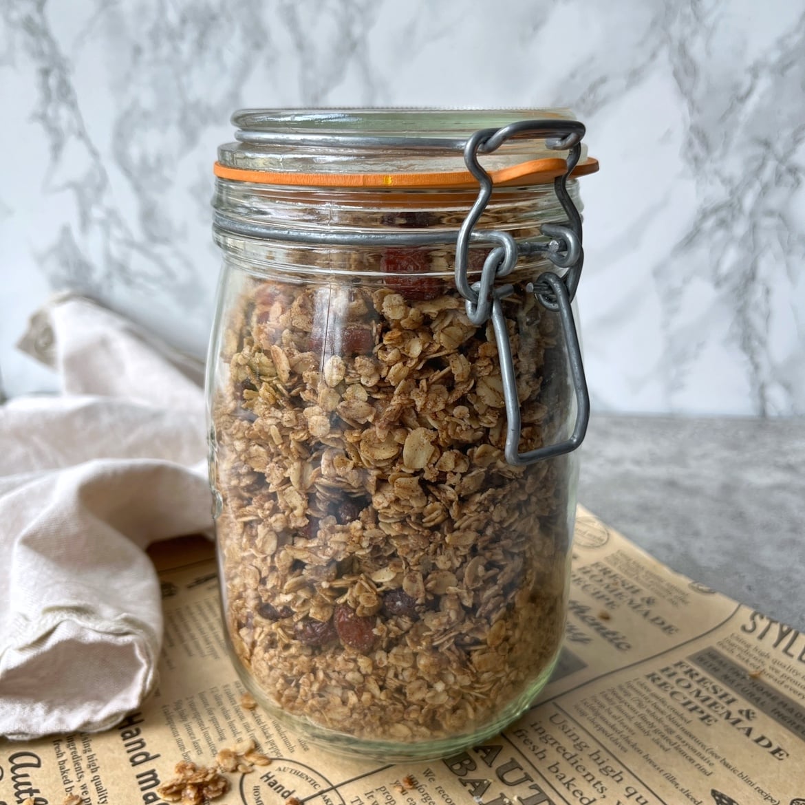 A glass jar filled with cinnamon granola sits on a newspaper with a cloth napkin in the background.
