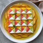 A beautiful geometric salad with quarters of watermelon, Galia melon, nectarine, and feta arranged on a white plate surrounded by a yellow vinaigrette.