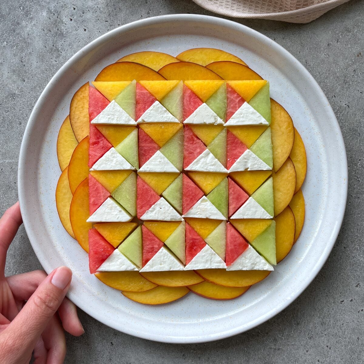 A hand holds a plate with a geometric salad featuring quarters of watermelon, Galia melon, nectarine, and feta surrounded by a yellow vinaigrette.
