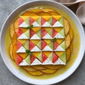A beautiful geometric salad with quarters of watermelon, Galia melon, nectarine, and feta arranged on a white plate surrounded by a yellow vinaigrette.