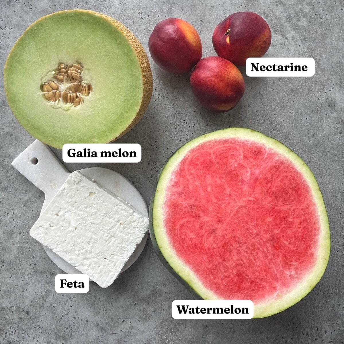 Ingredients for a Feta Cheese Salad featuring a half watermelon, Galia melon, three nectarines, and a block of feta on a grey surface.