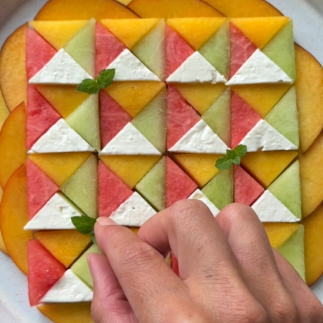 A hand places a fresh mint leaf on colourful melon, nectarine, and feta salad arranged in a geometric pattern.