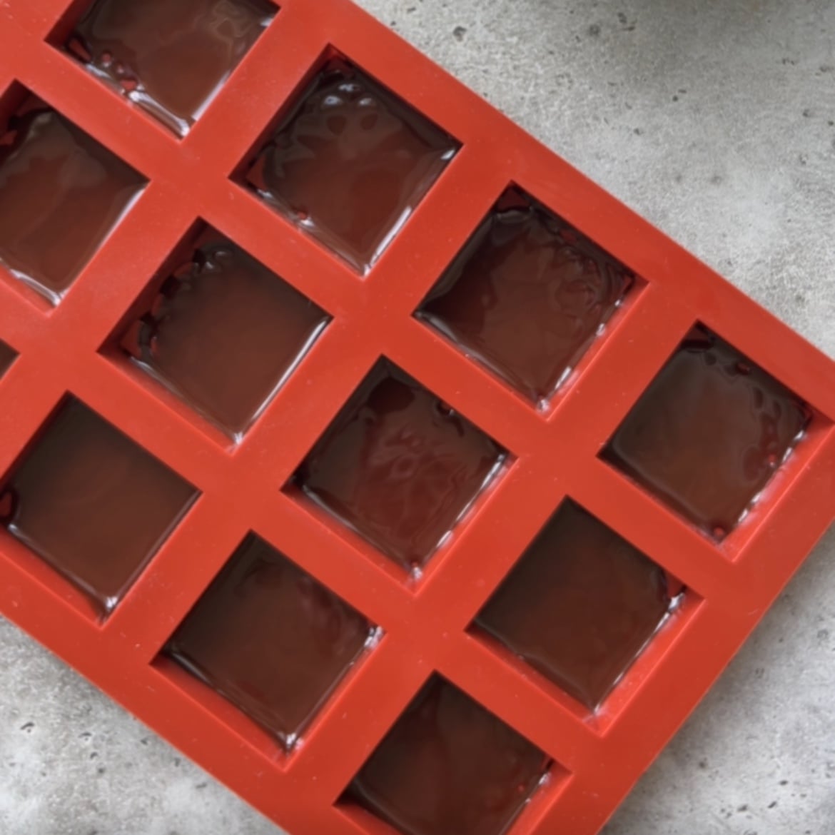 A red silicone mold with square cavities is filled with melted chocolate.