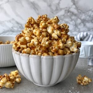 A white bowl filled with Homemade Caramel Popcorn sits on a gray surface, with a smaller dish and loose, golden popcorn pieces in the background.