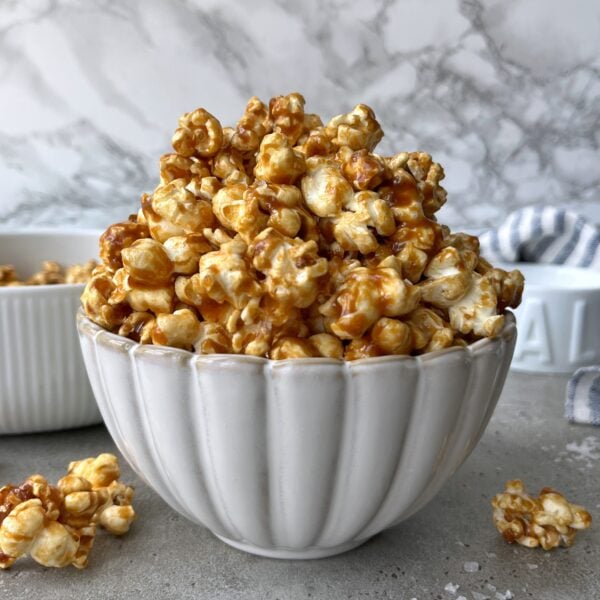 A white bowl filled with Homemade Caramel Popcorn sits on a gray surface, with a smaller dish and loose, golden popcorn pieces in the background.