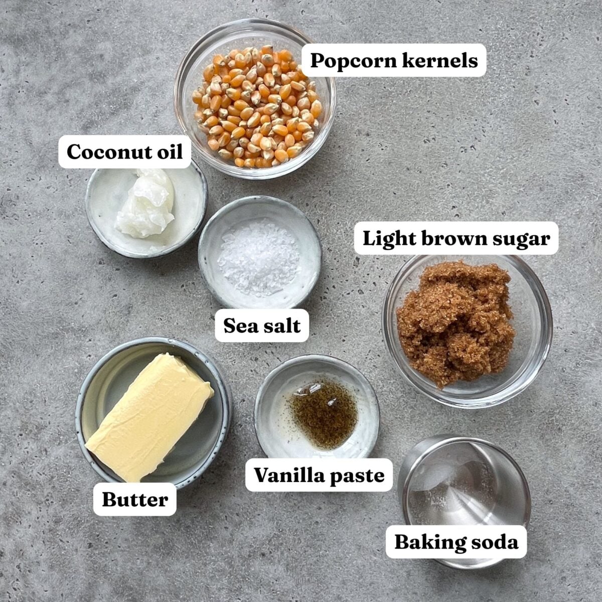 Seven small bowls containing the ingredients for Homemade Caramel Popcorn—popcorn kernels, coconut oil, sea salt, light brown sugar, butter, vanilla paste, and baking soda—sit labeled on a gray surface.
