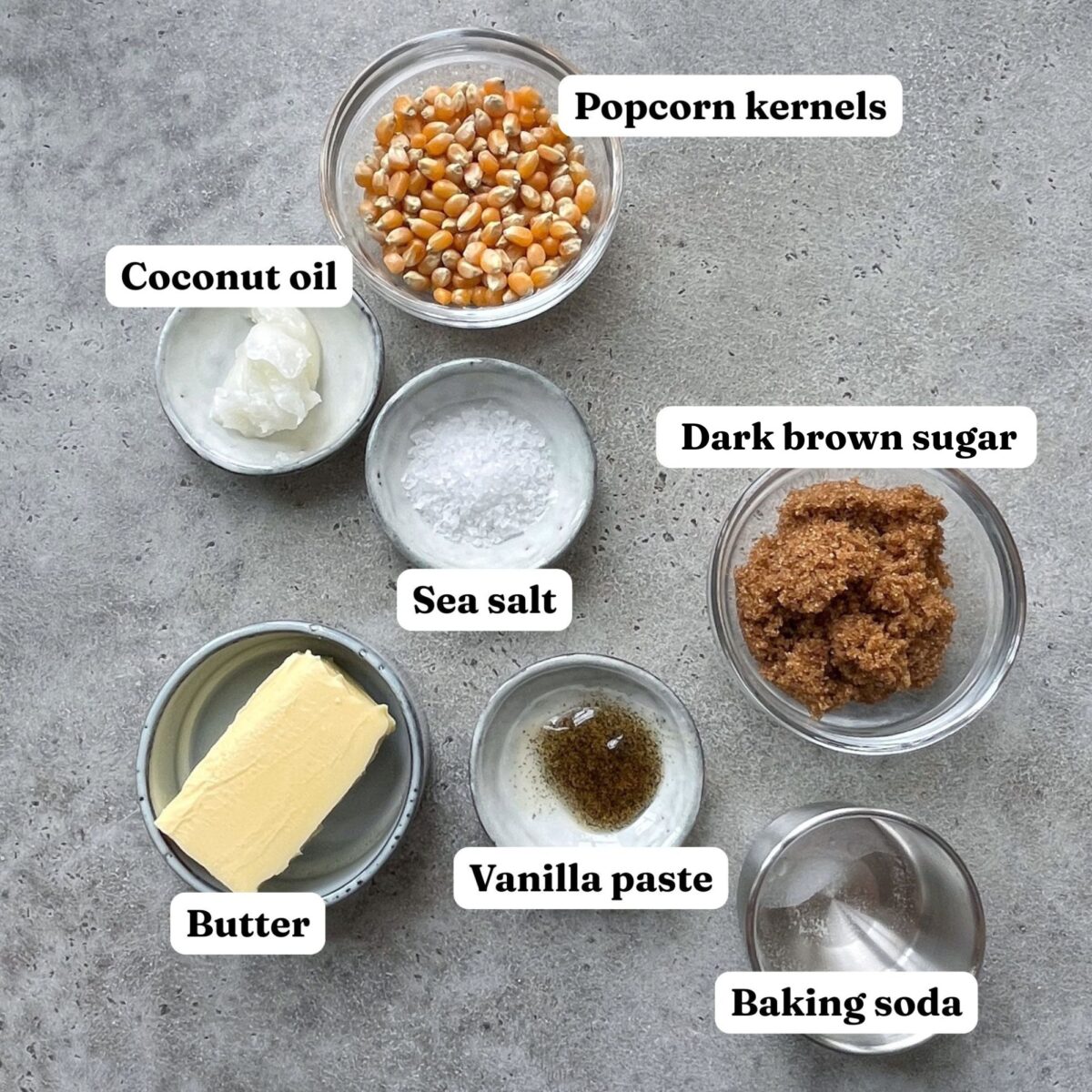 Seven small bowls featuring popcorn kernels, coconut oil, sea salt, dark brown sugar, butter, vanilla paste, and baking soda sit labeled on a gray surface.