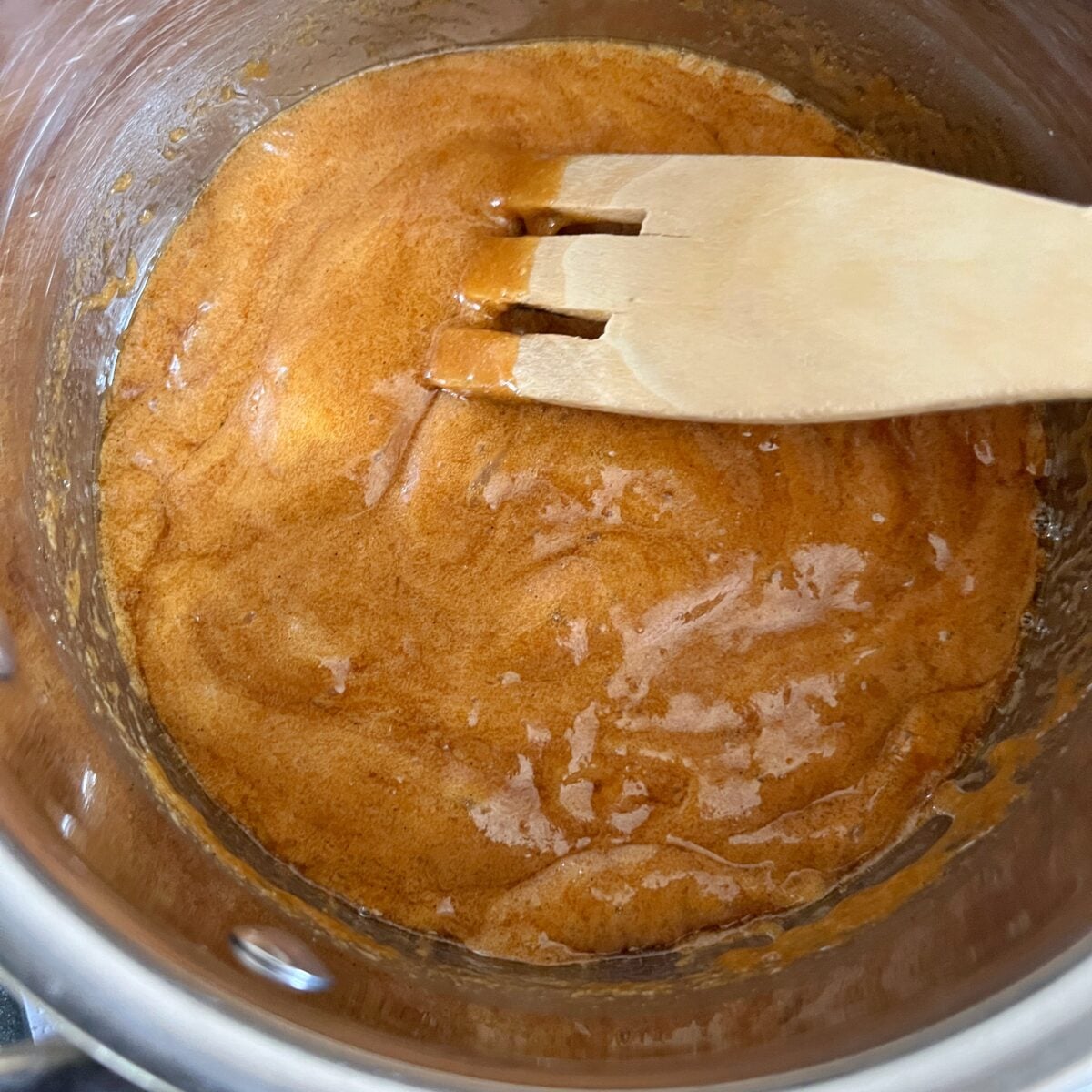A wooden spatula stirs baking soda into a thick, golden-brown caramel in a metal saucepan.