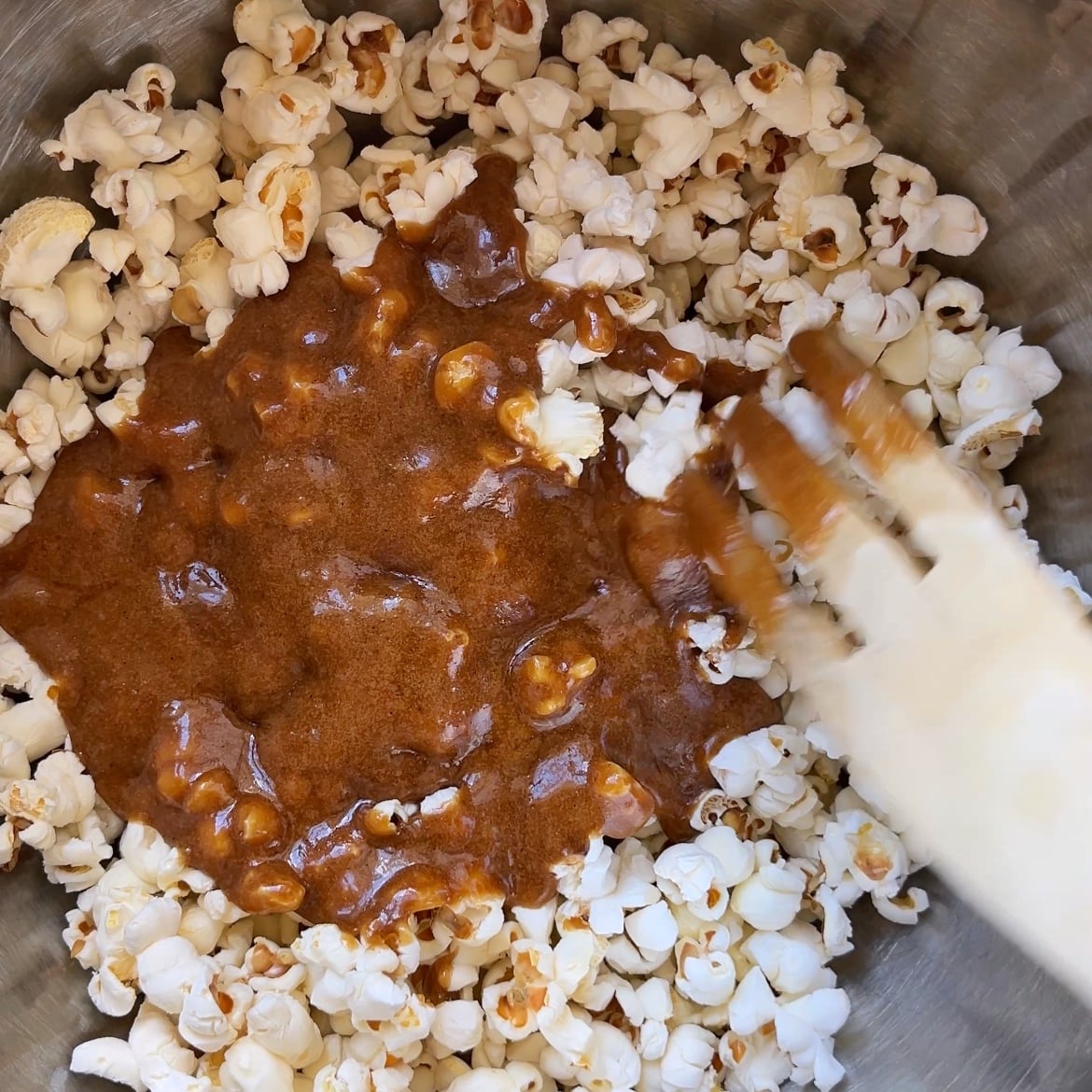 Homemade Caramel Popcorn being mixed in a metal bowl as a wooden utensil coats each piece with thick brown caramel.