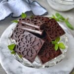 A glass dish holds homemade chocolate candy squares with a creamy white filling, garnished with fresh mint leaves, on a marble surface.