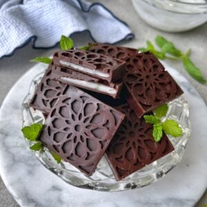 A glass dish holds homemade chocolate candy squares with a creamy white filling, garnished with fresh mint leaves, on a marble surface.