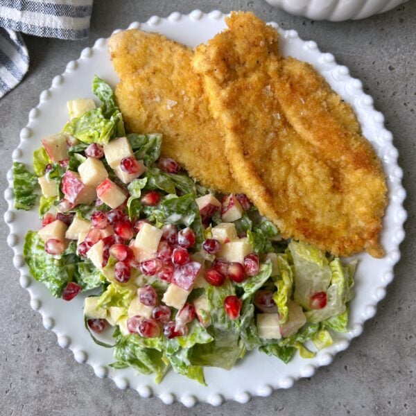 A white plate with two breaded, fried Milanese Chicken fillets next to a salad with lettuce, apple cubes, pomegranate seeds, and creamy dressing.