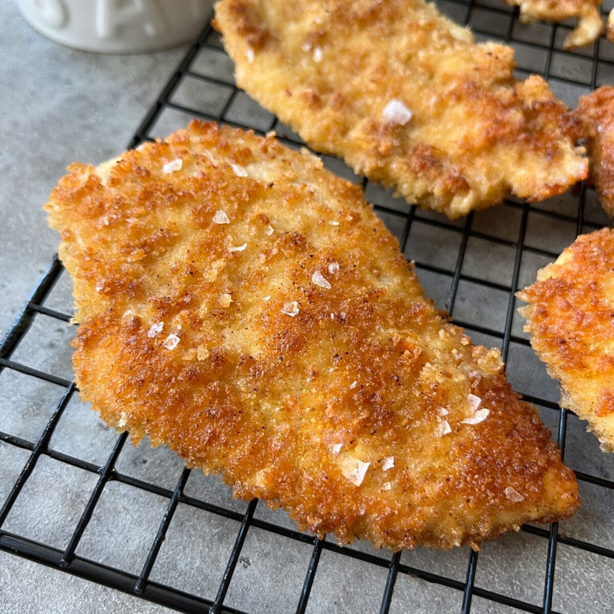 Breaded and fried Milanese Chicken cutlets cooling on a black wire rack, topped with coarse salt.