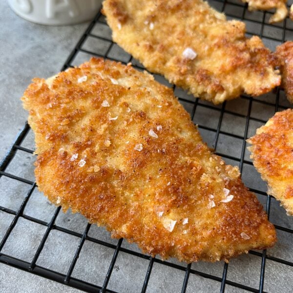 Breaded and fried Milanese Chicken cutlets cooling on a black wire rack, topped with coarse salt.