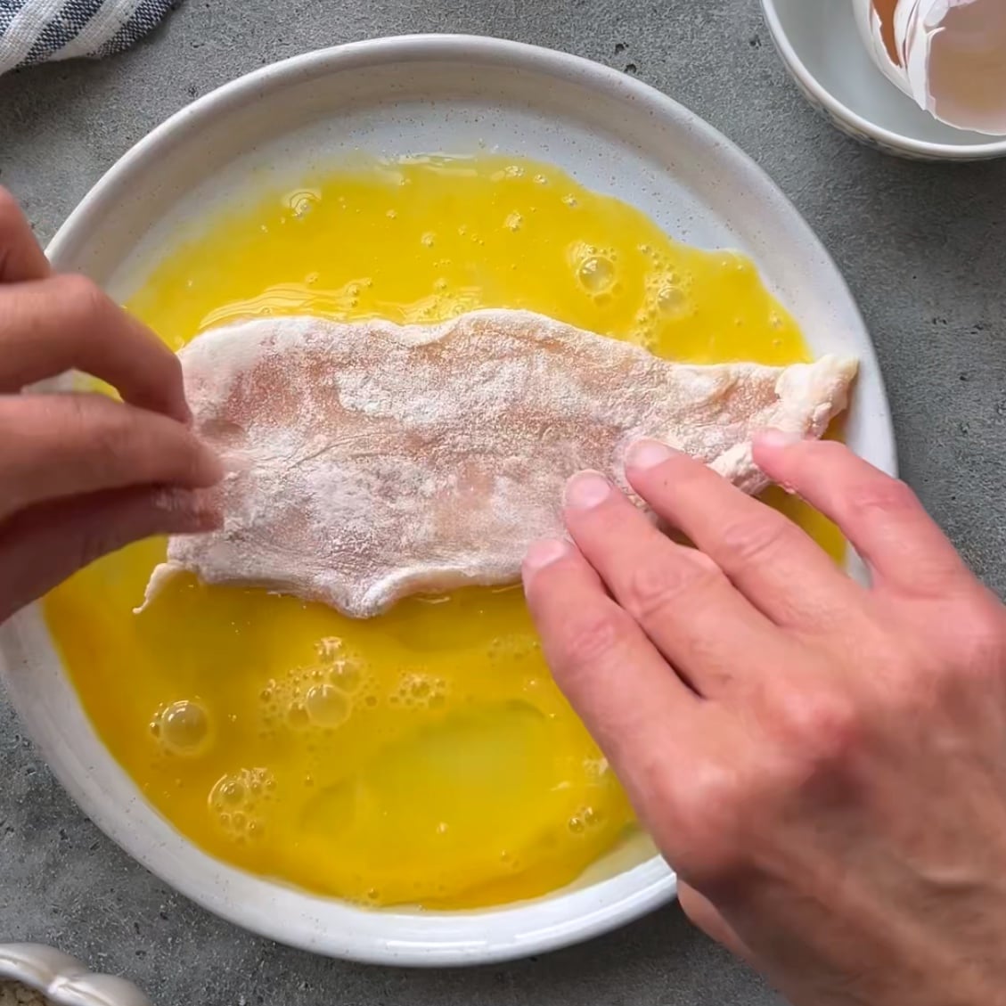 A person dips a raw, flour-coated chicken fillet into a plate of beaten eggs.