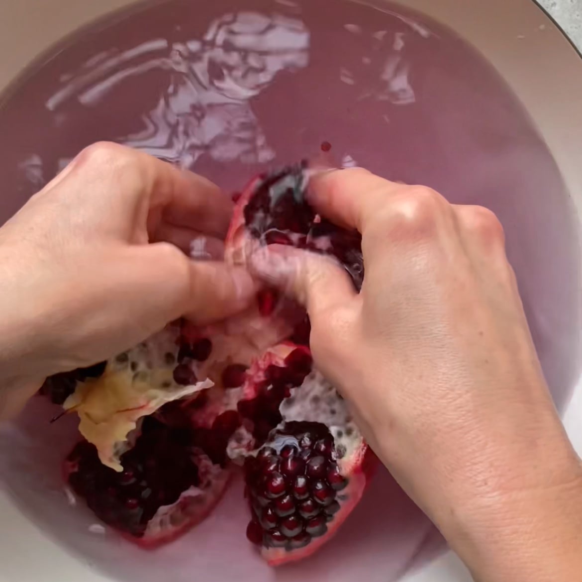 Two hands break apart a pomegranate underwater in a bowl.