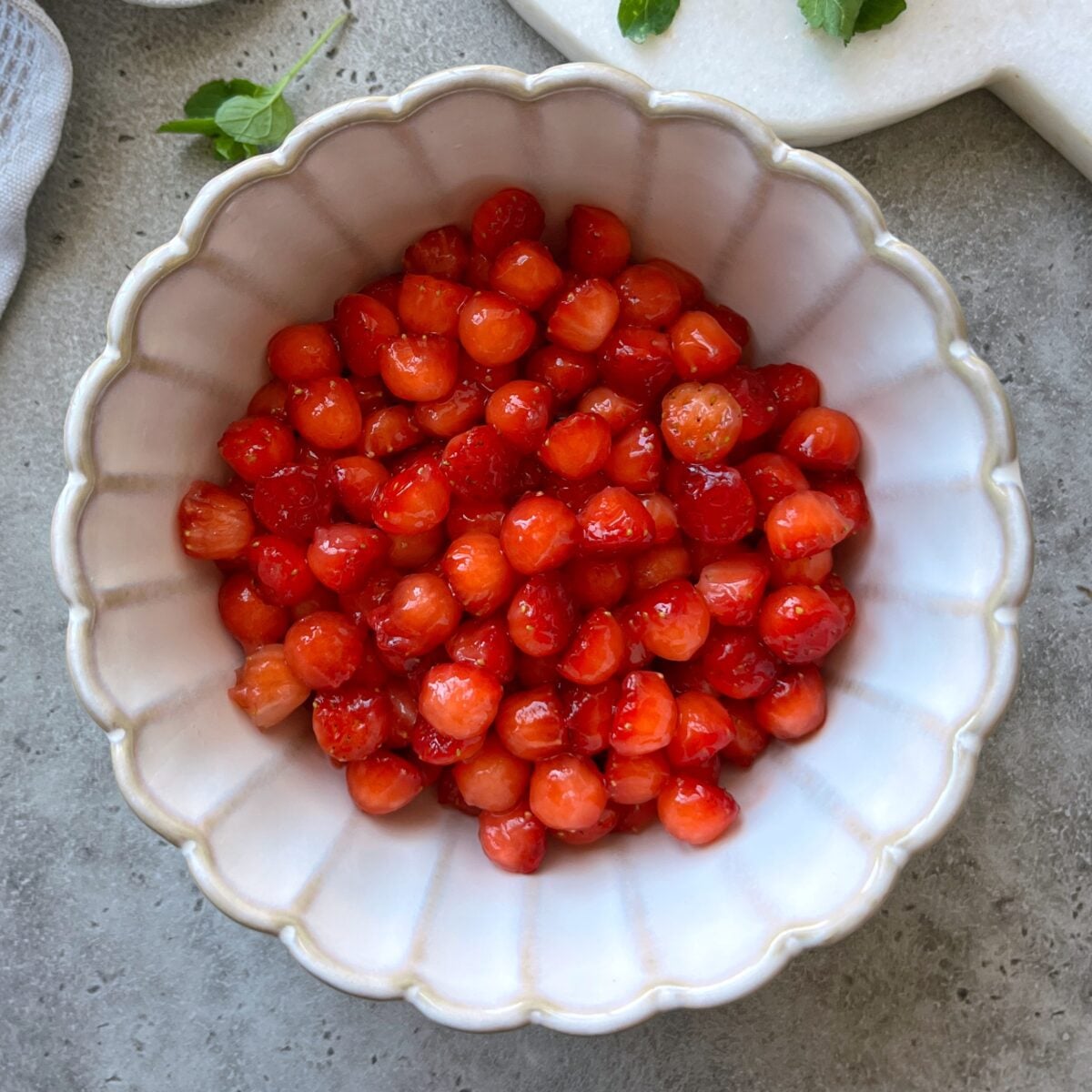 A white scalloped bowl filled with strawberry balls on a gray countertop.