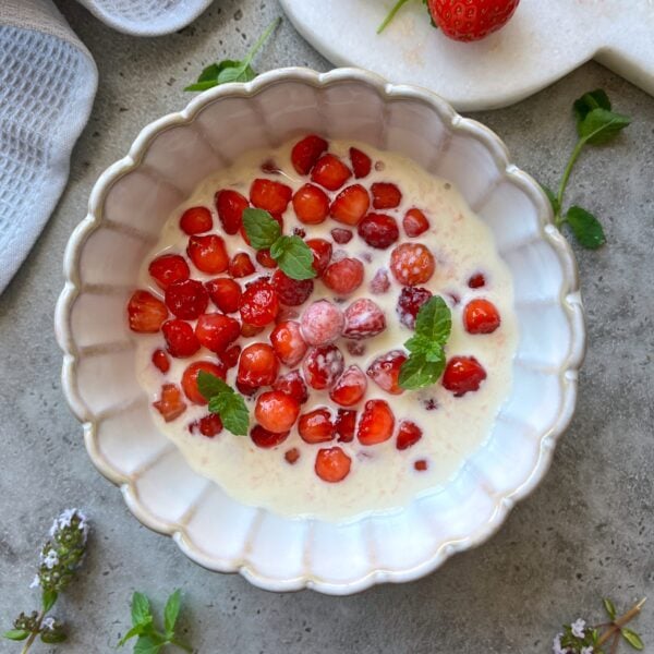 A white bowl filled with luscious strawberries and cream, garnished with fresh mint leaves, sits elegantly on a gray surface.