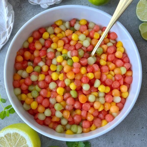 A white bowl filled with colorful watermelon, apricot, Galia melon, mango, and kiwi mini balls. Lime halves and a white cloth are nearby on a gray surface.
