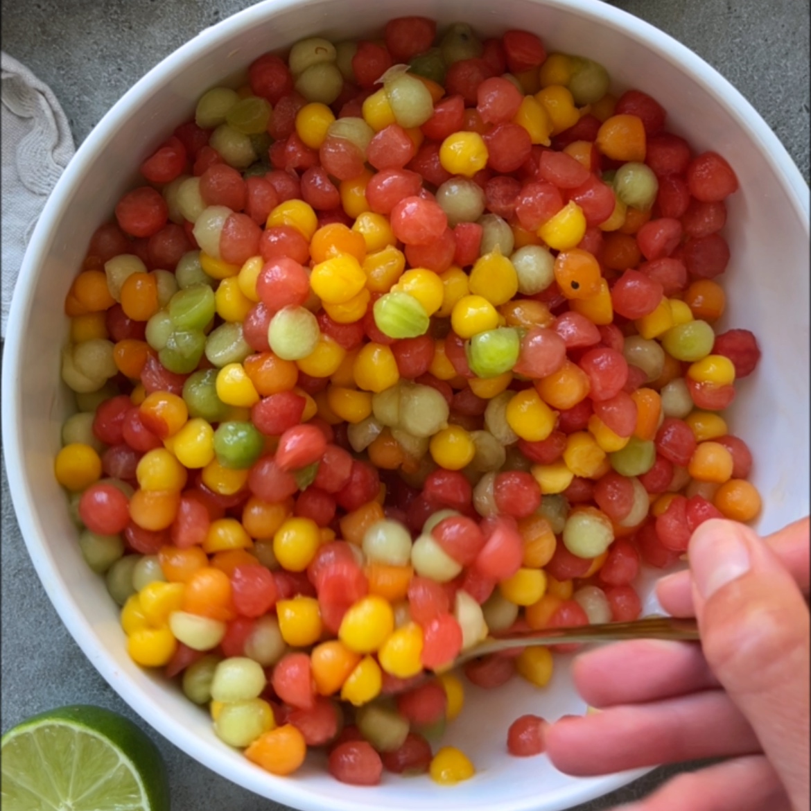 A hand mixes a bowl filled with small, round fruit balls.
