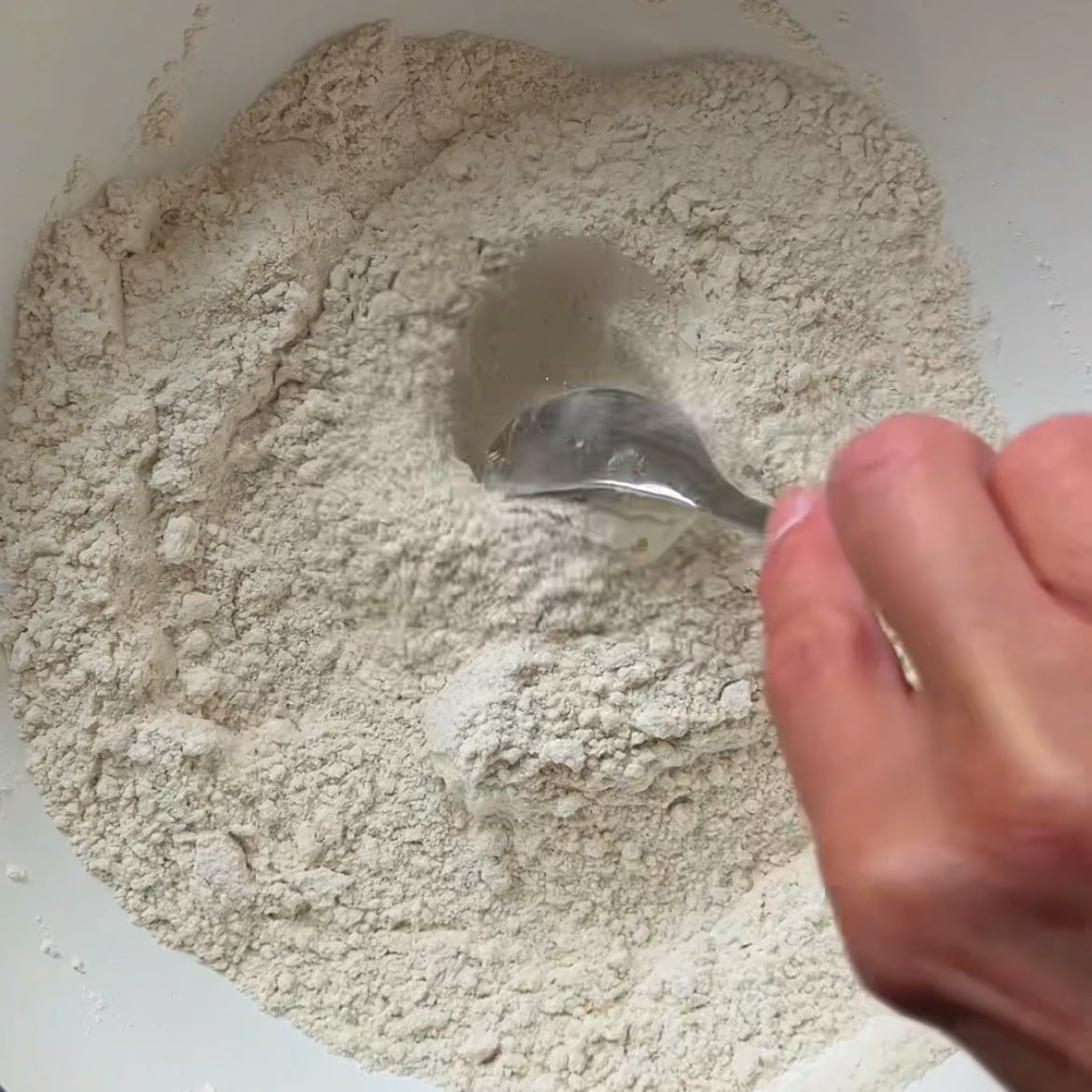 A hand mixes flour, sugar, and baking powder in a medium bowl.