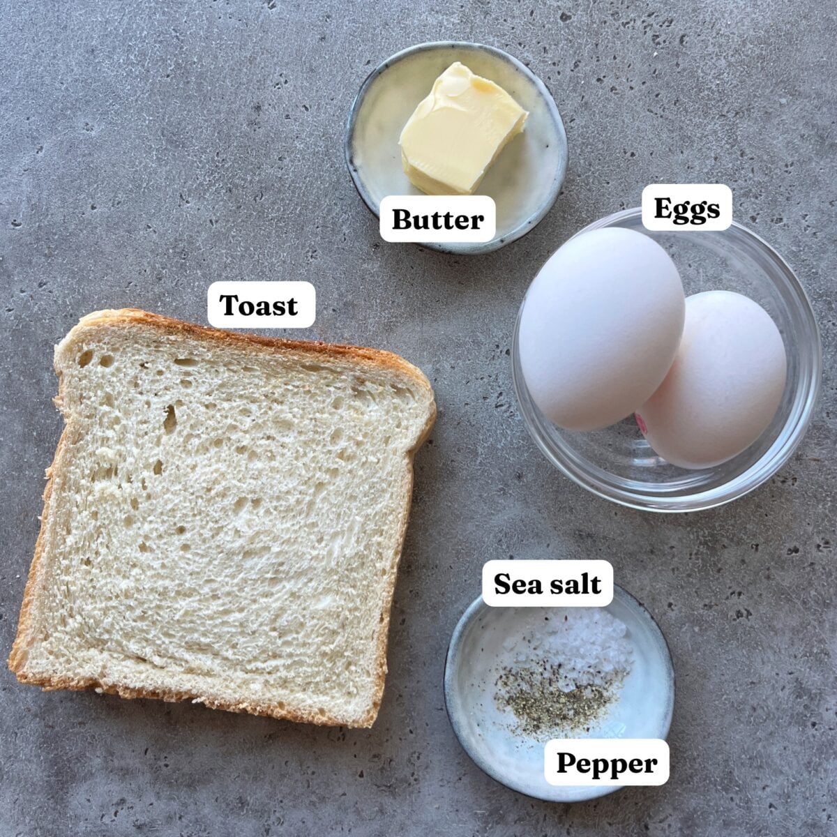 Ingredients for eggs in a hole, with two pieces of toast, butter, two eggs, and a bowl of sea salt, and pepper.