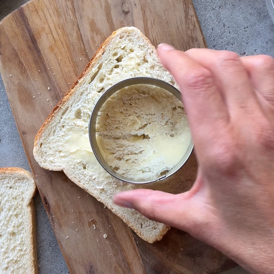 A hand uses a round cookie cutter to cut out a hole from a slice of buttered toast.