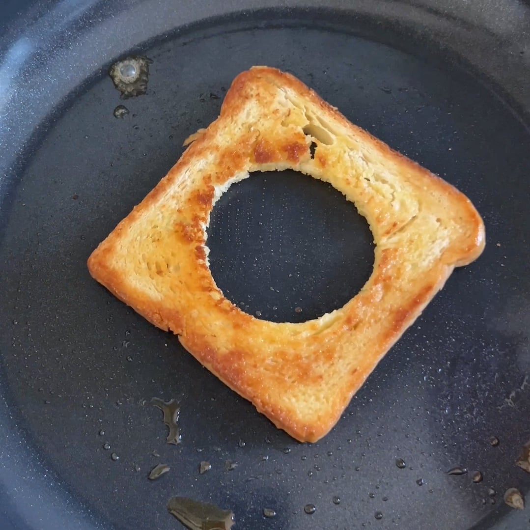 A piece of golden brown toast with a hole in it is frying in a pan.