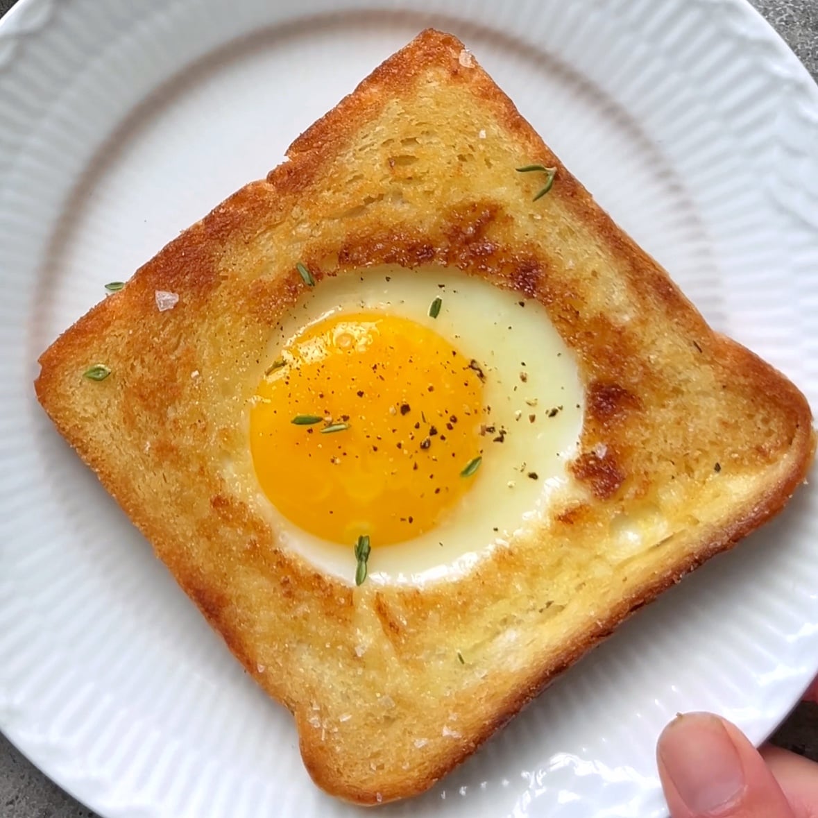 A white plate is topped with a freshly made golden-brown toast with a cooked egg in the center. The yolk is runny.