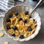 A scalloped bowl of mini pancakes topped with milk, maple syrup, and blueberries with a striped dish cloth and bowl of berries in the background.