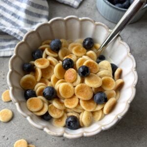 A scalloped bowl of mini pancakes topped with milk, maple syrup, and blueberries with a striped dish cloth and bowl of berries in the background.