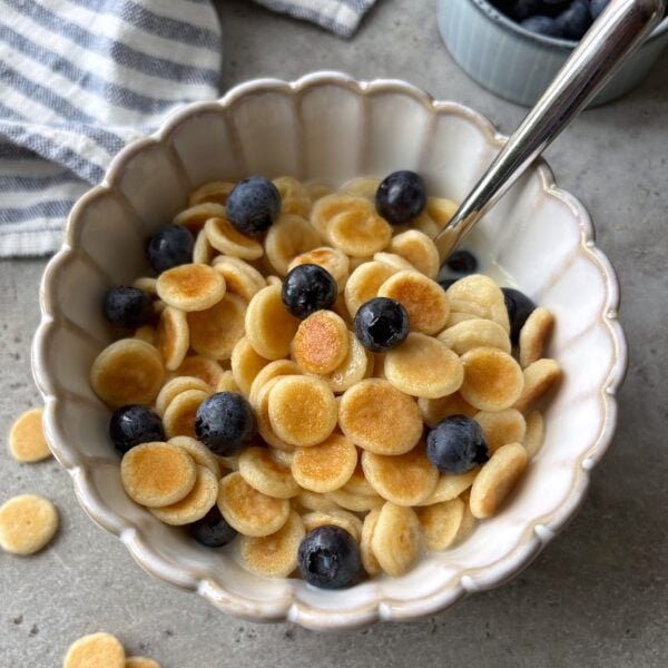 A scalloped bowl of mini pancakes topped with milk, maple syrup, and blueberries with a striped dish cloth and bowl of berries in the background.