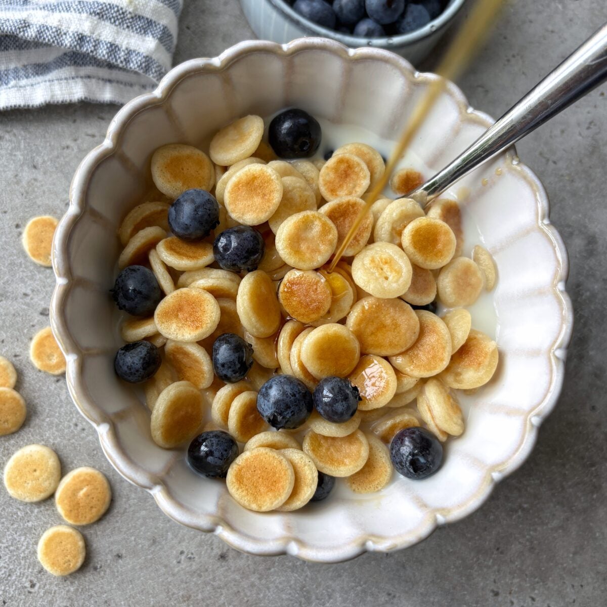 Maple syrup being poured onto a bowl of mini pancake cereal topped with blueberries and milk.