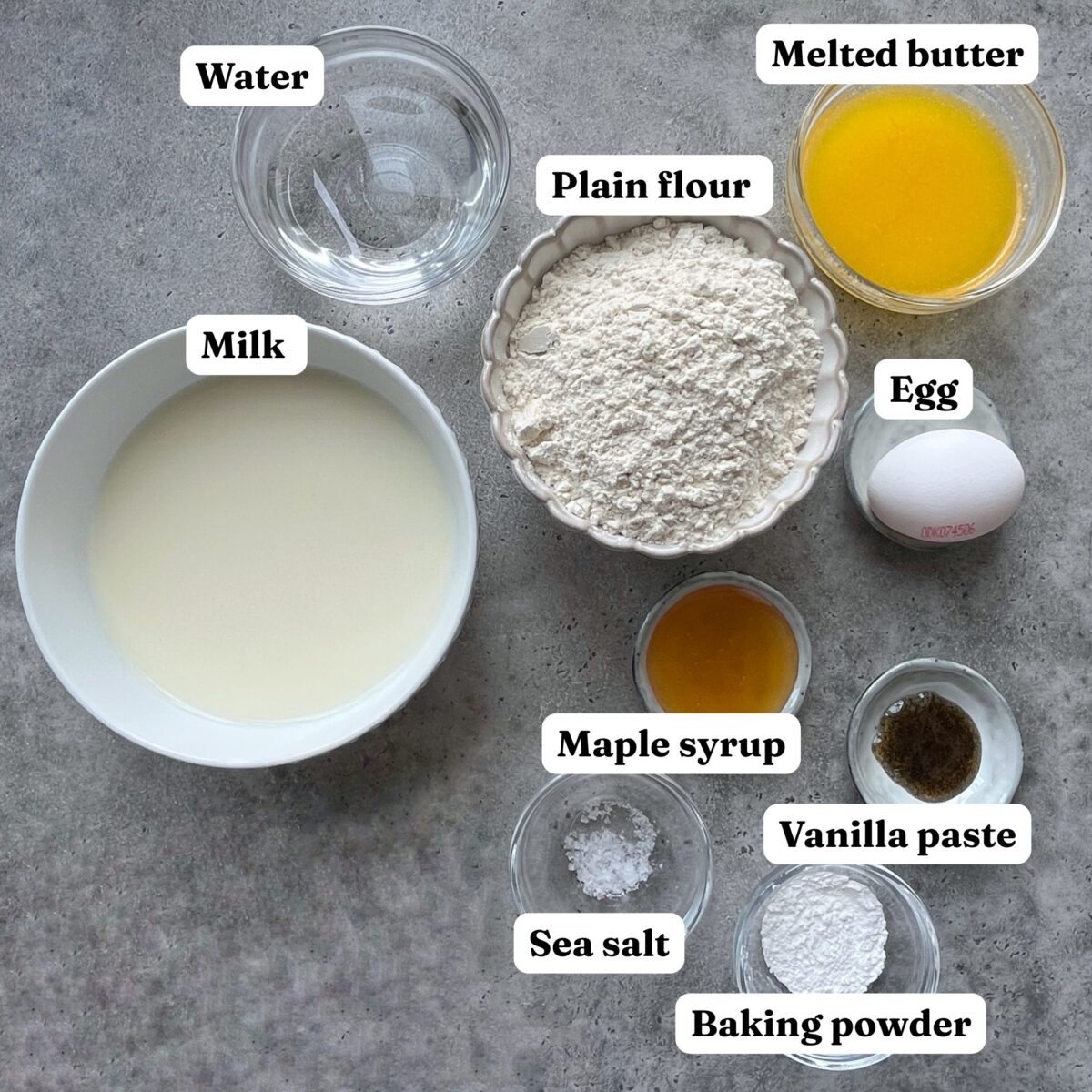 A bowl of milk, water, melted butter, plain flour, egg, maple syrup, vanilla paste, sea salt, and baking powder on a gray surface.