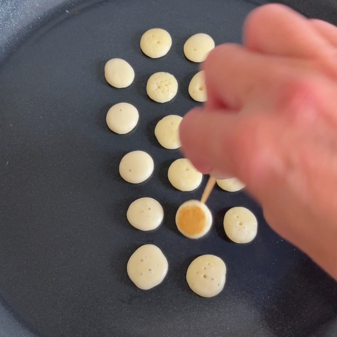 A hand uses a toothpick to turn over mini pancakes on a hot pan.
