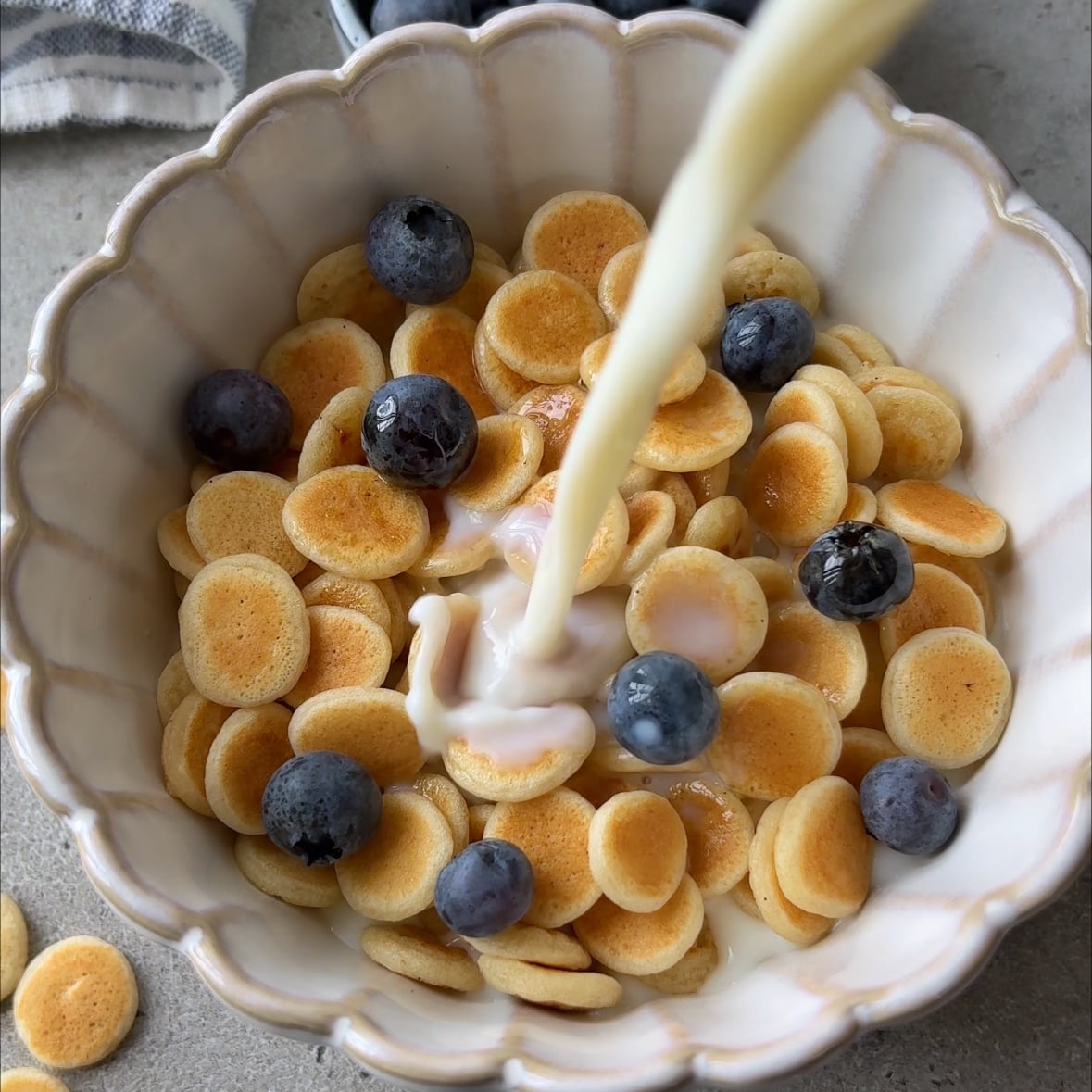 Mini pancakes in a bowl topped with maple syrup and blueberries. A hand pours milk on top.